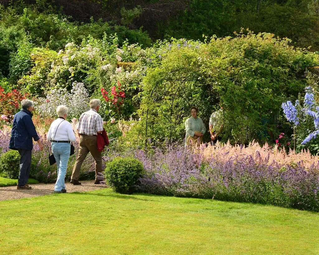 Een groep van vier mensen loopt over een tuinpad naar een met groen begroeid hekwerk, waar een andere persoon staat. De weelderige privétuinen van Oost-Schotland staan vol kleurrijke bloemen en dichte planten.