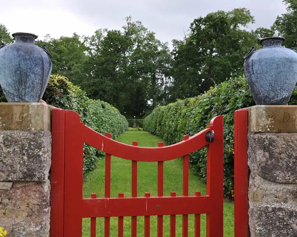 Een felrood houten hek staat tussen twee stenen pilaren met daarop grote blauwe keramieken vazen en geeft toegang tot keurig gesnoeide privétuinen in Oost-Schotland, met op de achtergrond een groen tuinpad omzoomd door heggen en bomen.