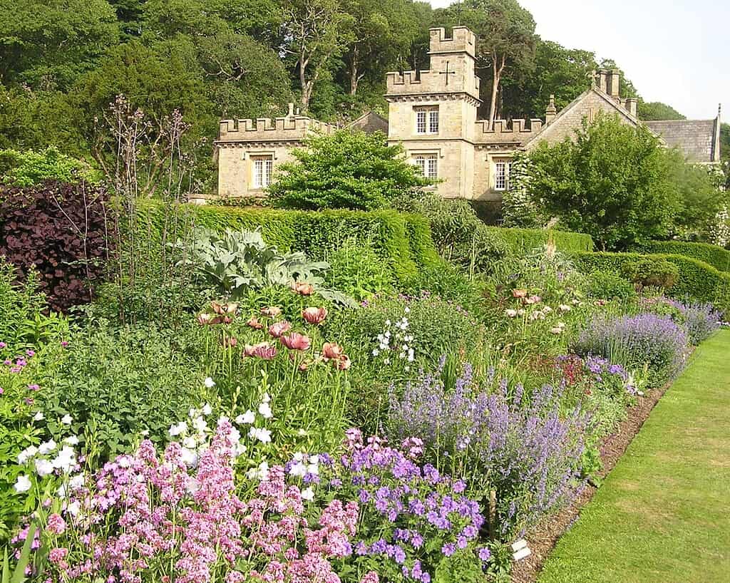 Een weelderige, kleurrijke tuin met bloeiende bloemen en struiken ligt voor een stenen herenhuis met gekanteelde torens, die de charme van de privétuinen van Oost-Schotland weerspiegelen, omringd door dichte groene bomen onder een heldere hemel.