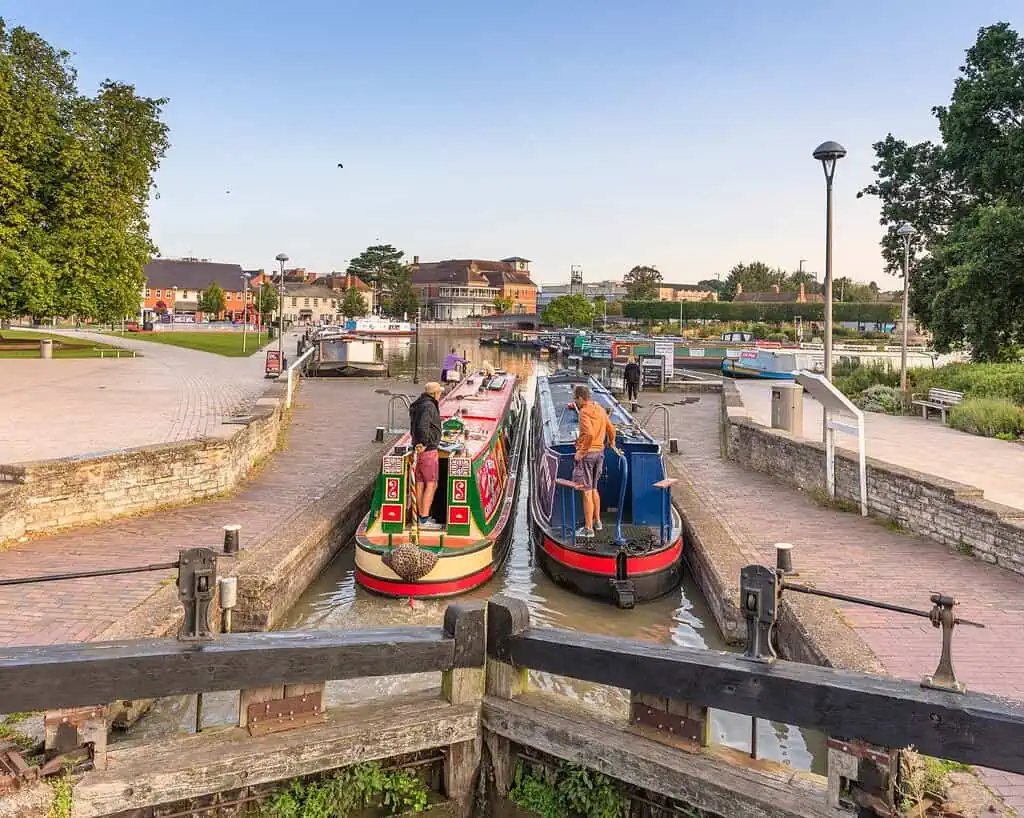 Twee kleurrijke kanaalboten varen door een sluis op een zonnige dag in een klein Brits stadje, met mensen op de boten en schilderachtige landhuizen, bomen en bankjes zichtbaar op de achtergrond.