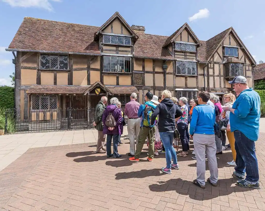 Een groep toeristen staat op een zonnige dag voor een historisch vakwerkhuis met glas-in-loodramen en een pannendak, dat doet denken aan de tijd van Shakespeare. Sommigen bewonderen het gebouw, terwijl anderen een praatje maken in de tuin, die doet denken aan klassieke tuinen en landhuizen.