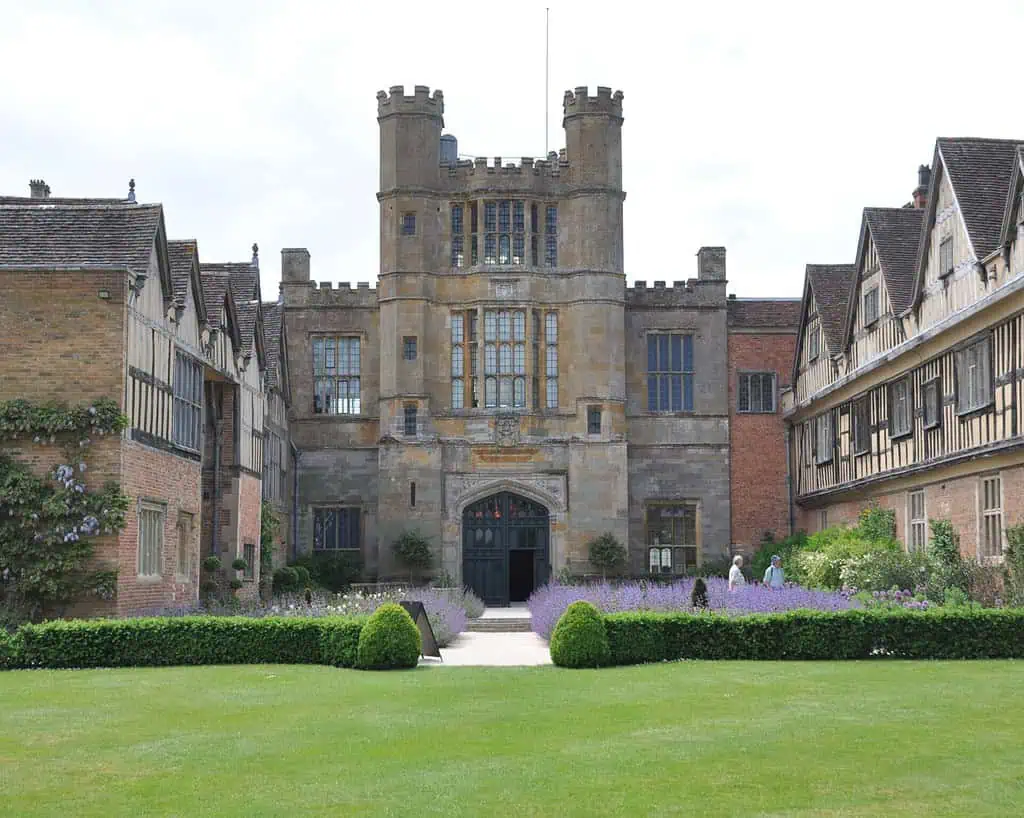 Kijk door een raam van een historisch bakstenen herenhuis dat doet denken aan de tijd van Shakespeare, met grote, sierlijke ramen, decoratief steenwerk en weelderige tuinen aan de rand van de geplaveide binnenplaats.