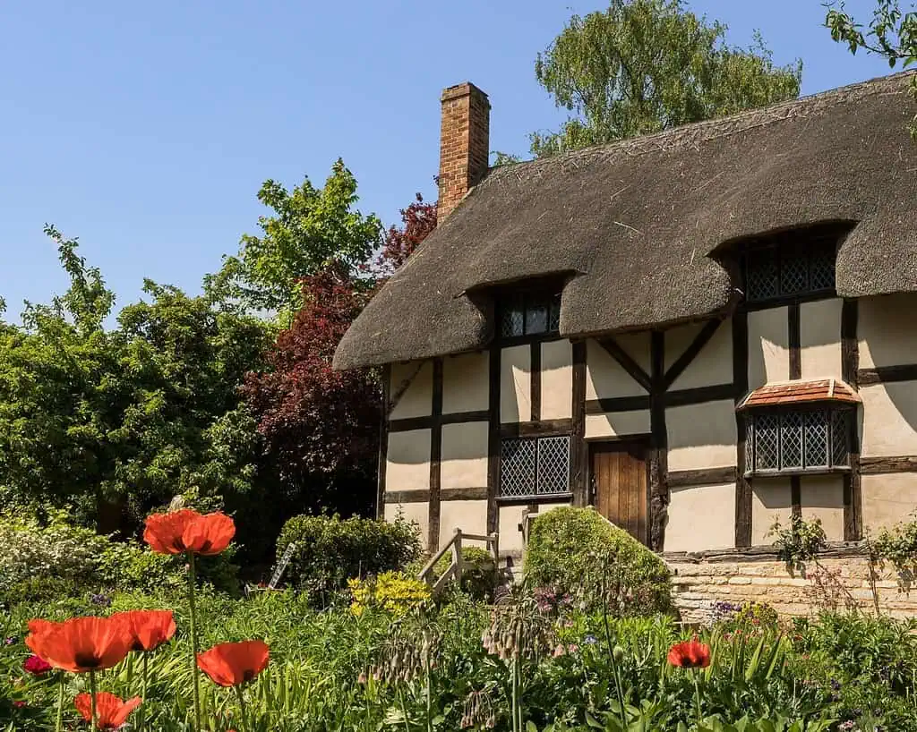 Een weelderige kamer, die doet denken aan grote landhuizen, heeft groen fluwelen fauteuils, sierlijke houten tafels, decoratieve lampen en vazen. De muren zijn gevuld met ingelijste schilderijen en portretten onder een uitgebreide kroonluchter boven een gedessineerd tapijt.