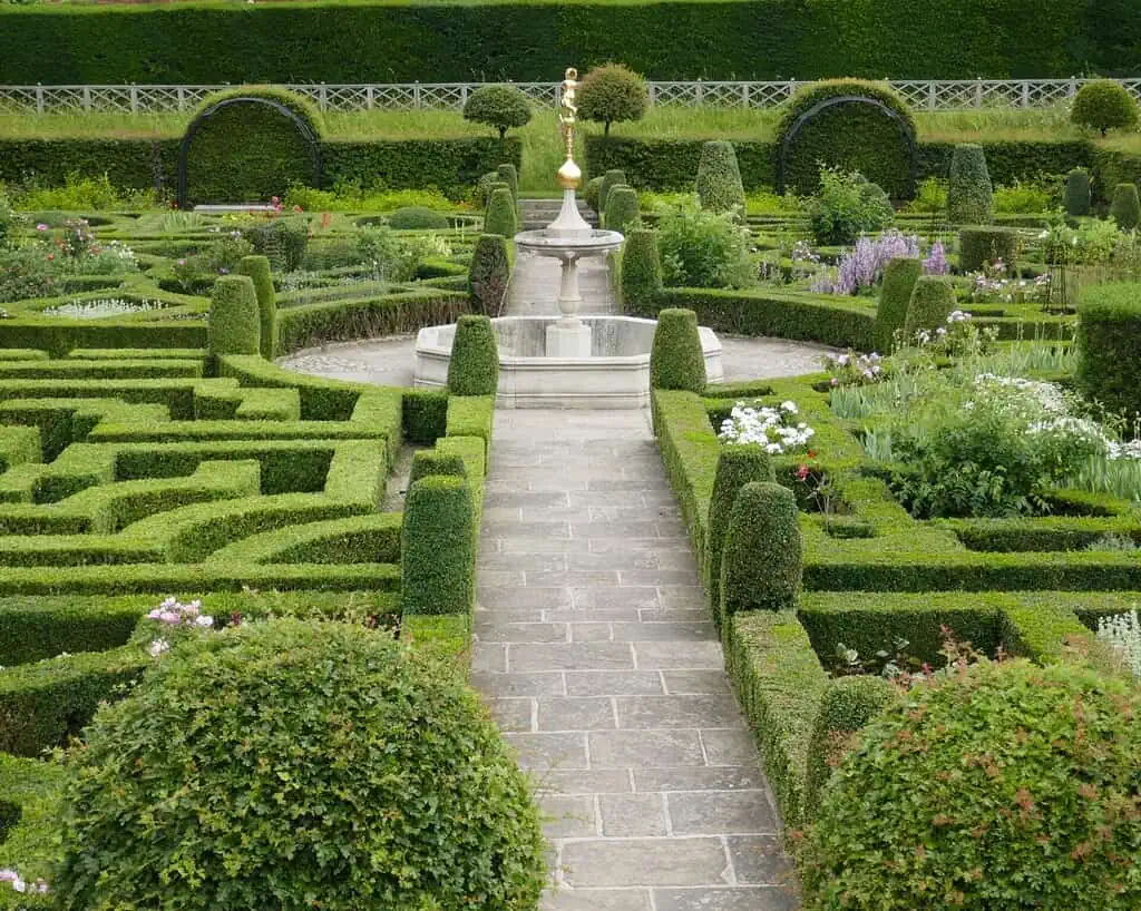 Een formele tuin die doet denken aan landhuizen, met keurig gesnoeide groene heggen die een doolhof vormen, stenen paden, kleurrijke bloemen en een centrale stenen fontein omringd door gemanicuurde tuinen en elegante vormsnoei.