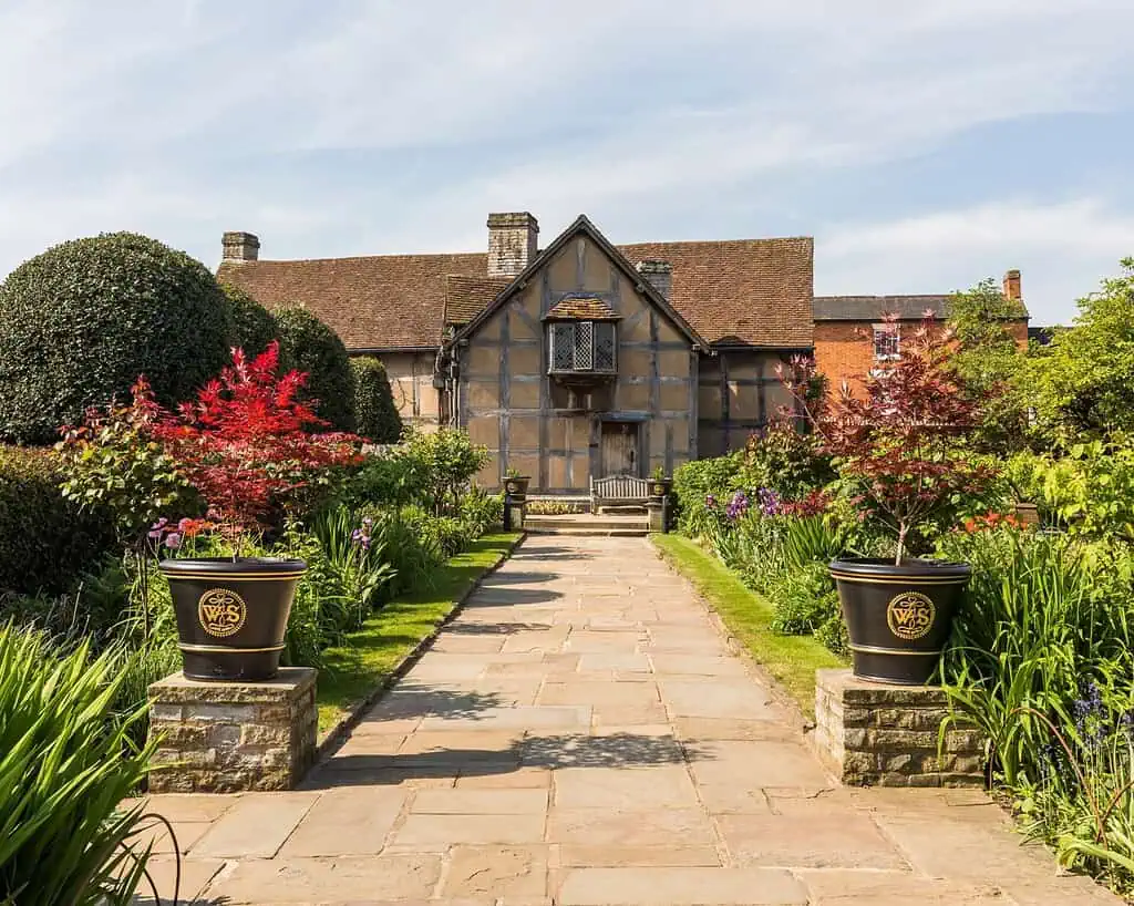 Een stenen pad leidt naar een historisch landhuis in vakwerkstijl met een zadeldak, omringd door gemanicuurde tuinen, levendige bloemen en twee grote potplanten bij de ingang onder een strakblauwe hemel.