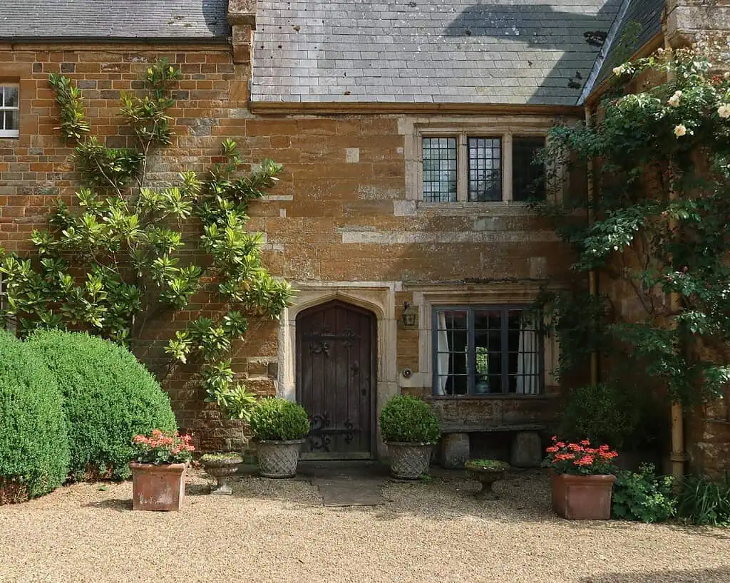 Een rustiek stenen huis in Shropshire met een houten deur, boogramen en weelderig groene planten die tegen de muren opklimmen. De binnenplaats van grind nodigt je uit om het rustig aan te doen tussen levendige rode bloemen in klassieke plantenbakken.