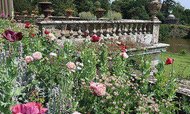 Een levendige bloementuin met rode en roze bloemen groeit naast een oude stenen balustrade in Shropshire, met uitzicht op een weelderig groen landschap en een vijver onder een helderblauwe hemel - perfect om het rustig aan te doen.
