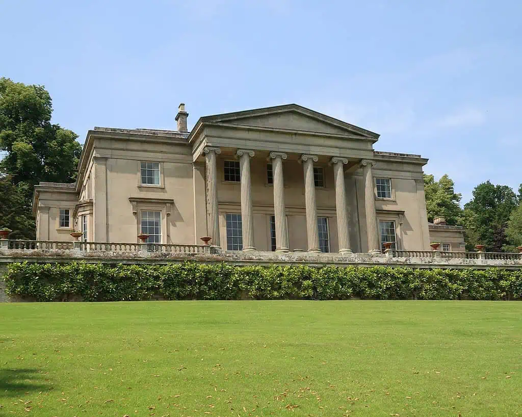 Een groot neoklassiek herenhuis in Shropshire, met hoge zuilen bij de ingang, grote ramen en een gemanicuurd gazon - perfect om het rustig aan te doen te midden van weelderig groen onder een heldere hemel.