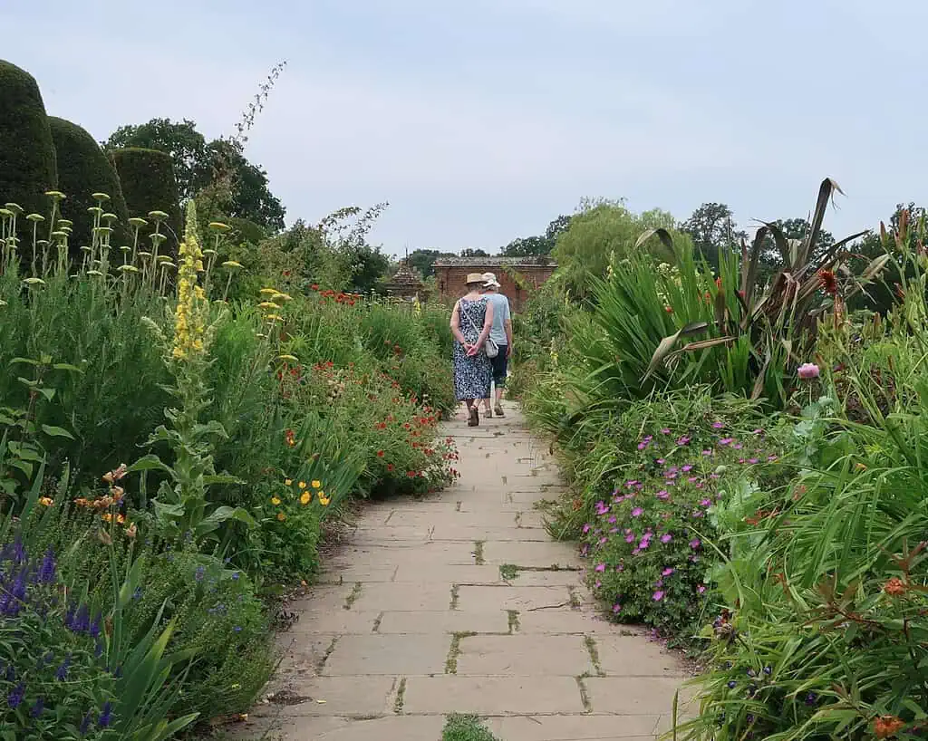 Twee mensen met zomerhoeden doen het rustig aan terwijl ze op een heldere dag over een stenen pad wandelen omringd door hoge, kleurrijke bloemen en weelderige groene planten in een tuin in Shropshire.