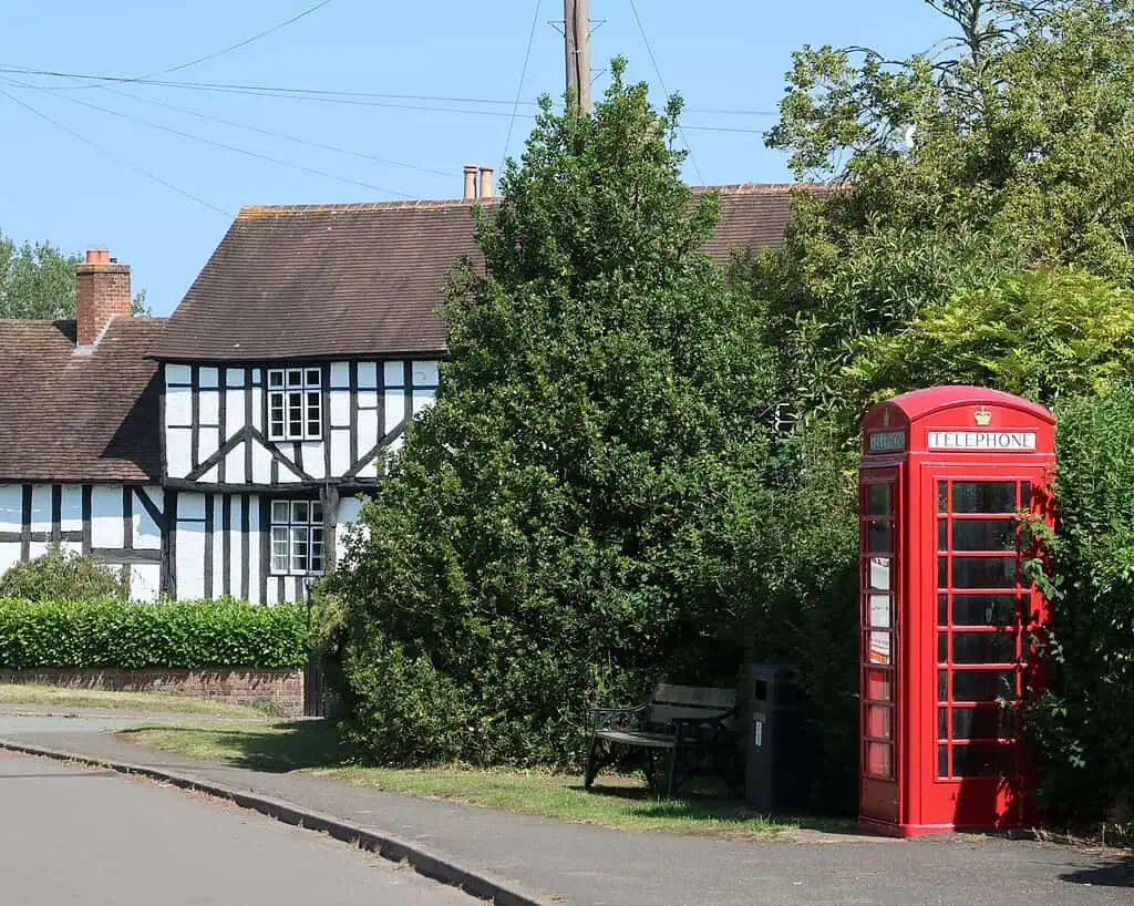 Een traditionele rode Britse telefooncel staat bij een bankje en weelderige groene struiken in Shropshire, met een vakwerkhuisje en heggen op de achtergrond - perfect om het rustig aan te doen op een zonnige dag.