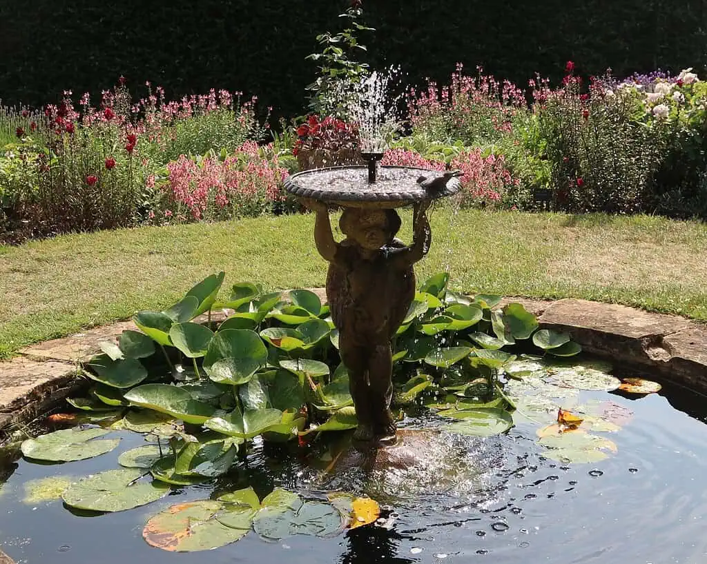 Een kleine stenen fontein in de vorm van een kind dat een vogelbadje vasthoudt staat in het midden van een vijver met lelieblad, omringd door bloeiende bloemen en groen - perfect om het rustig aan te doen in een vredige tuin in Shropshire.