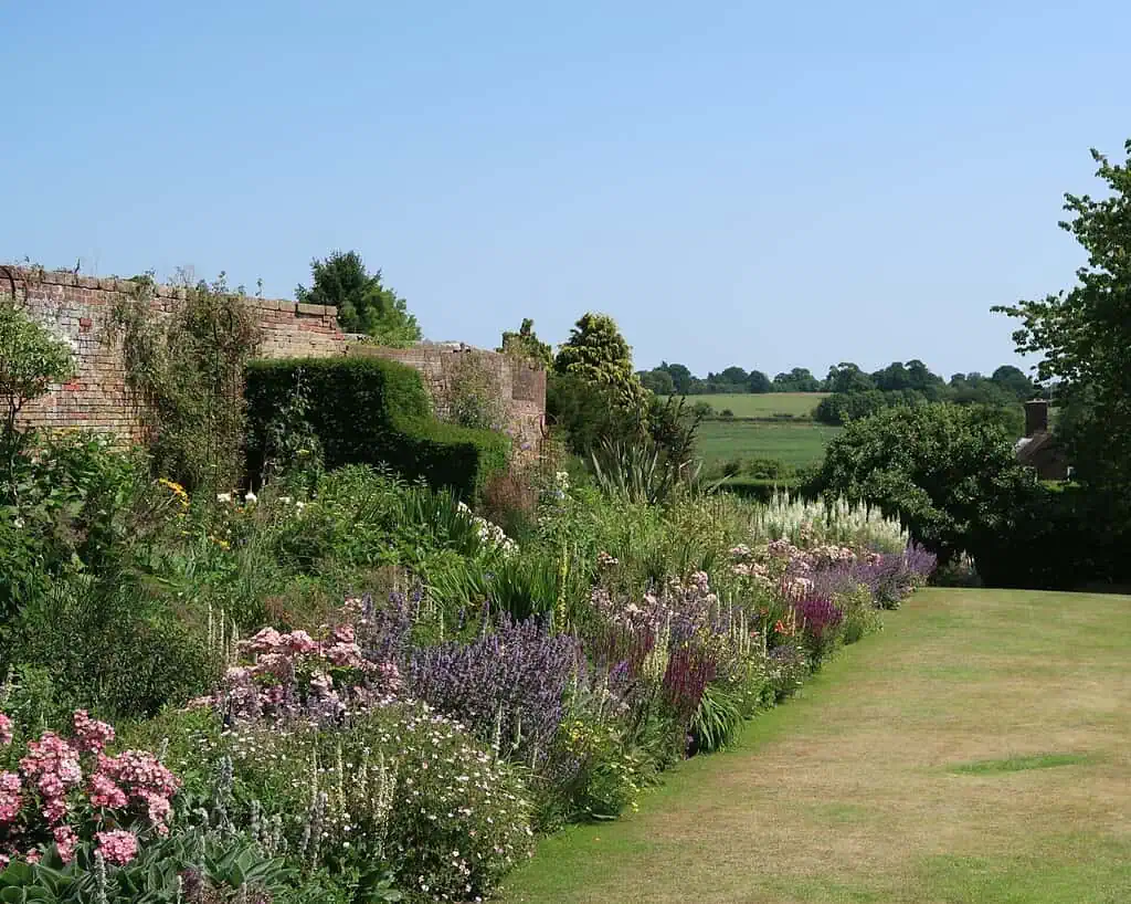 Een weelderige tuin in Shropshire met kleurrijke bloemen en groene struiken omzoomt een bakstenen muur onder een helderblauwe hemel, met open velden en bomen op de achtergrond - perfect om het rustig aan te doen.