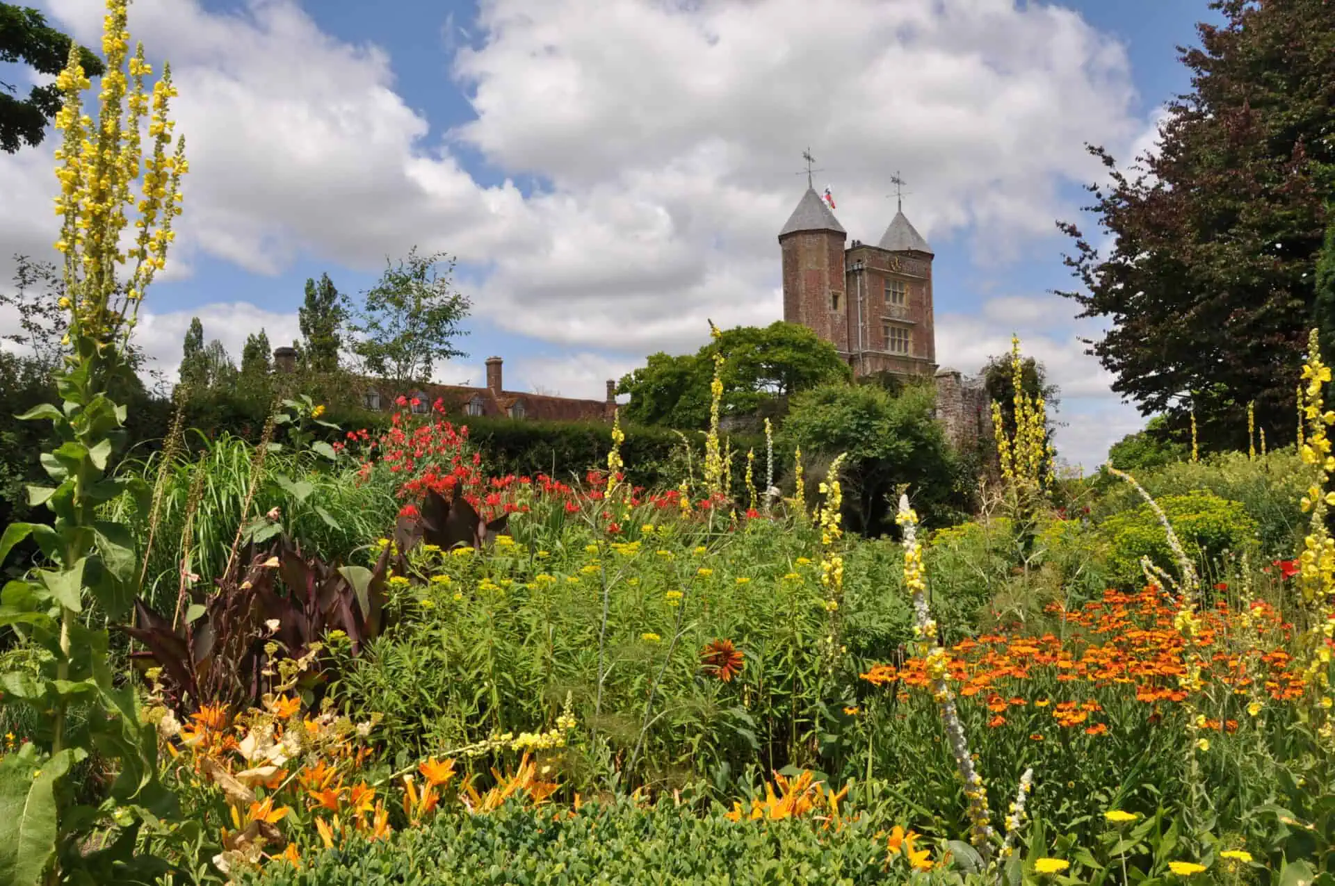 Een kleurrijke tuin vol gele, oranje en rode bloemen in volle bloei roept de sfeer op van de mooiste tuinen van Engeland, met op de achtergrond een historisch bakstenen kasteel en zijn twee hoge torens onder een gedeeltelijk bewolkte hemel.