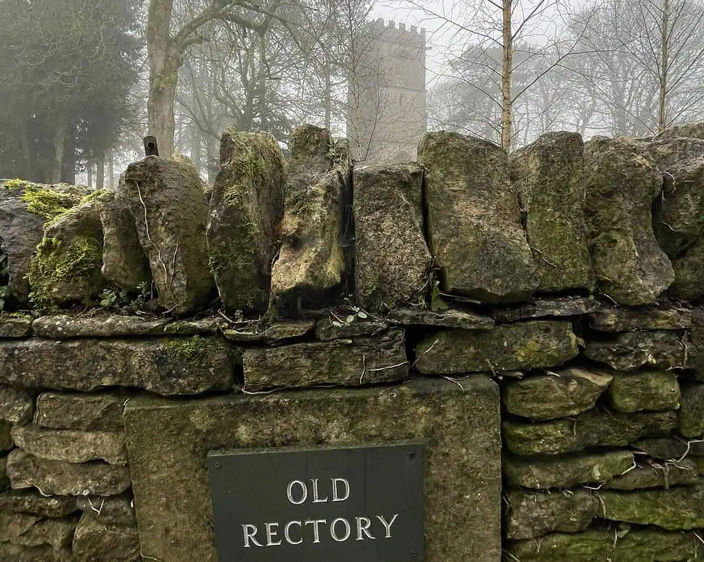 Een bemoste stenen muur met een gedenkplaat met de tekst Old Rectory staat voor bladerloze bomen en de hoge, vierkante stenen toren van Shaftesbury, gedeeltelijk zichtbaar in de mistige achtergrond - een rustig tafereel voor elke Sneeuwklokjesreis.