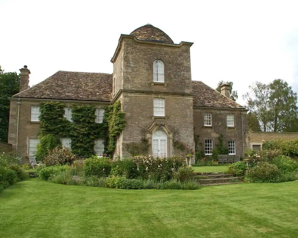 Een groot historisch stenen huis met klimop tegen de muren, omringd door weelderige groene gazons, kleurrijke bloembedden en een goed onderhouden tuin, onder een bewolkte hemel.