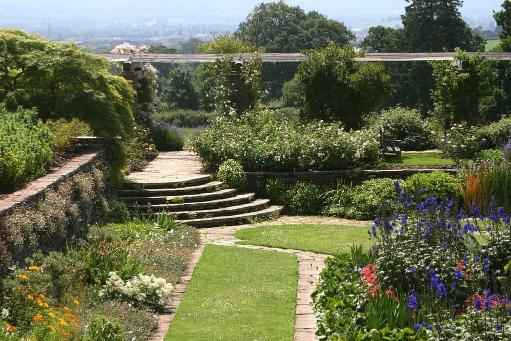 Een weelderige tuin met gebogen stenen trappen, kleurrijke bloemen, groene struiken en een stenen pad dat door goed onderhouden gazons en bloemperken leidt, tegen een achtergrond van verre bomen en heuvels.