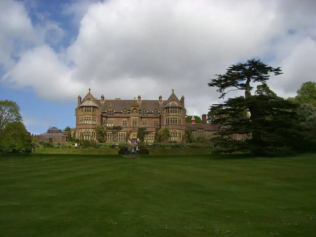 Een groot, historisch herenhuis met sierlijke architectuur ligt op een breed, verzorgd gazon onder een gedeeltelijk bewolkte hemel, met een hoge boom rechts en tuinbeelden ervoor.