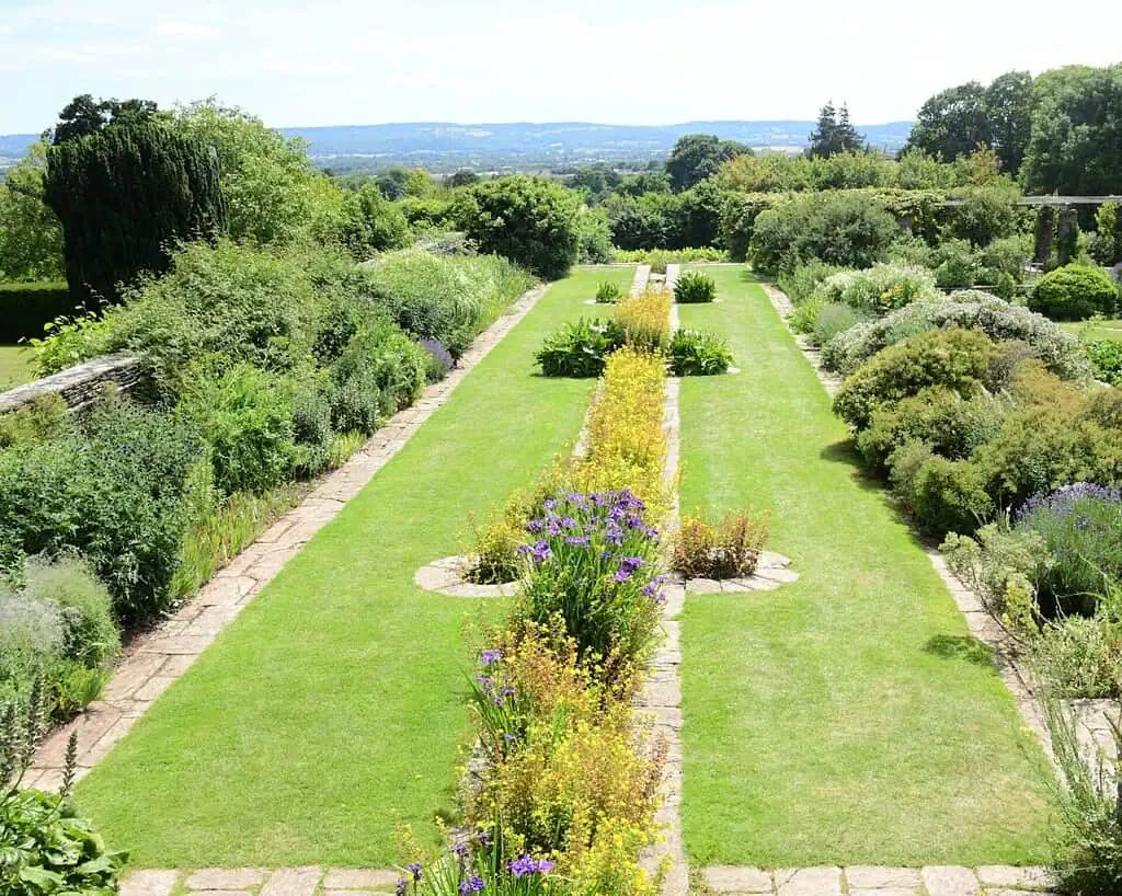 Een formele tuin met keurig gemaaide graspaden, bloemperken vol kleurrijke bloemen en struiken, en bomen en heuvels in de verte onder een heldere, halfbewolkte hemel.