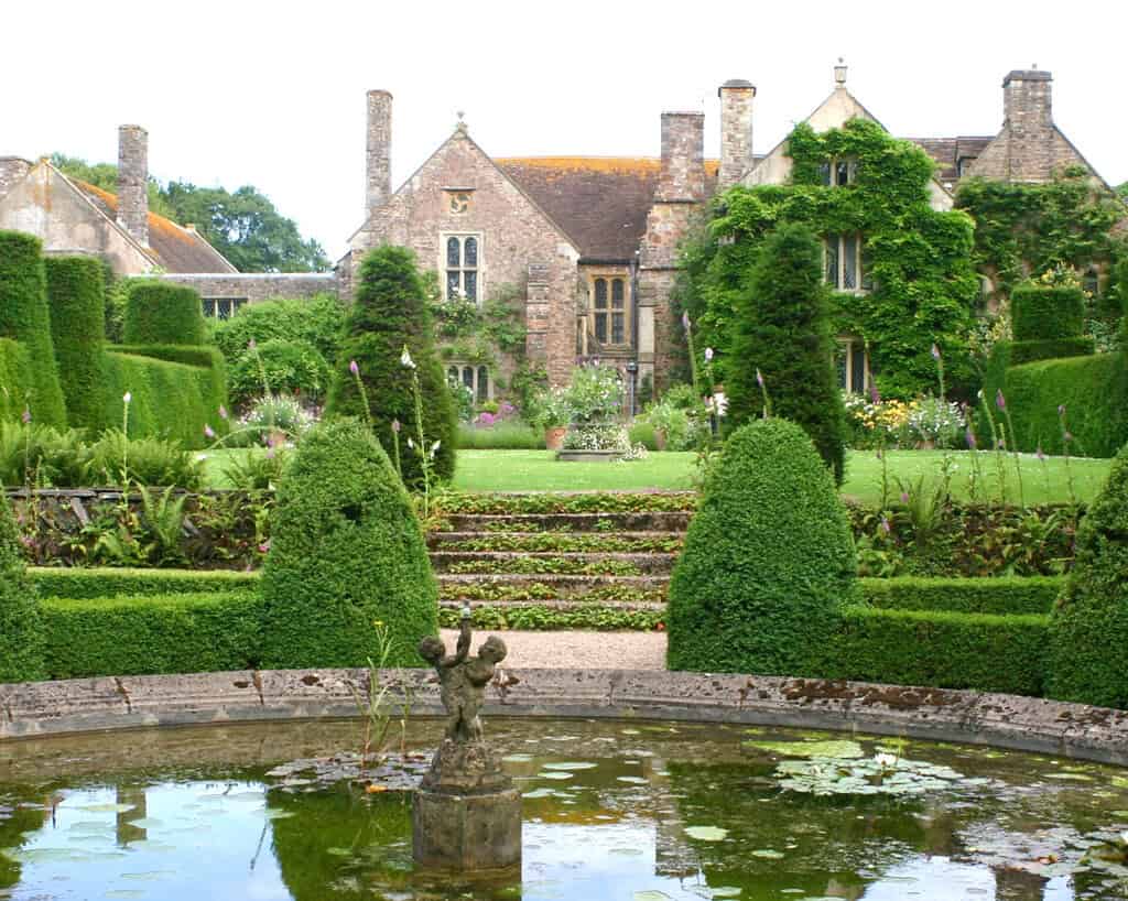 Een formele Engelse tuin met gemanicuurde heggen, een ronde vijver met een kleine fontein en een oud stenen landhuis bedekt met klimop op de achtergrond. Stenen trappen leiden van de vijver naar het huis.