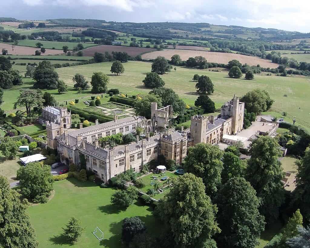 Luchtfoto van een groot historisch kasteel met stenen muren, omringd door verzorgde tuinen, gazons en bomen, in een landelijk landschap met glooiende velden en heuvels in de verte.
