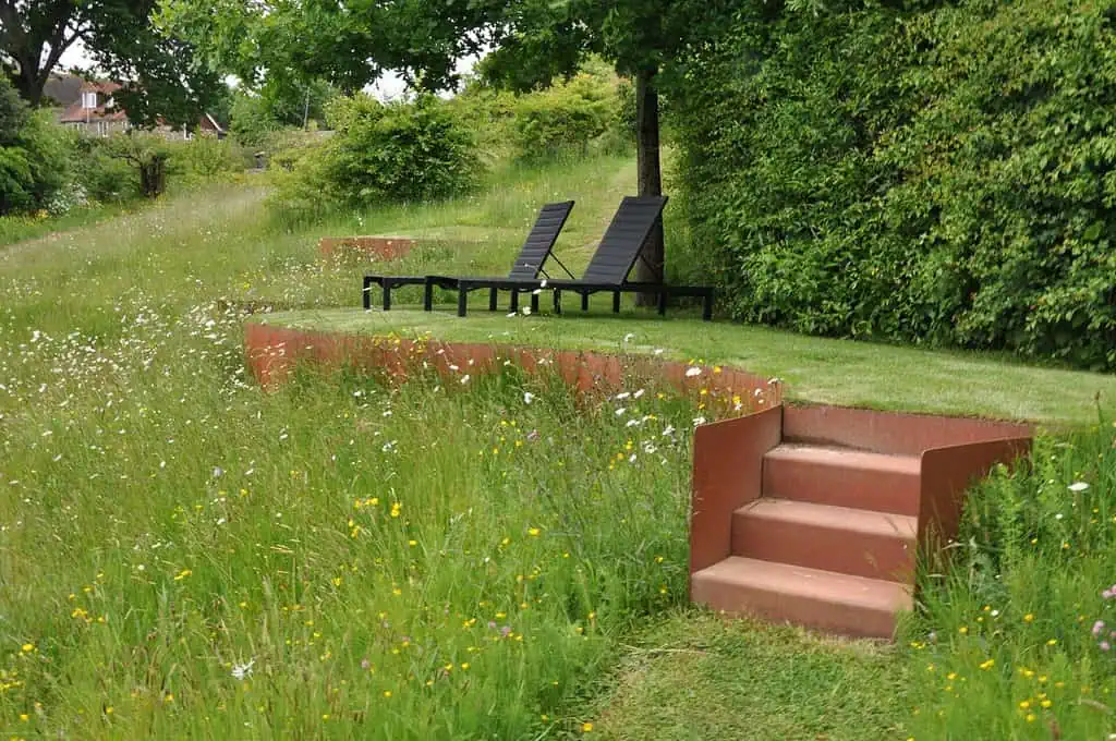 Twee zwarte loungestoelen staan op een platform van gras, omzoomd door weelderige hagen en wilde bloemen. Een rode trap leidt naar deze serene zithoek, een prachtig voorbeeld van moderne tuinen in een vredige tuin.