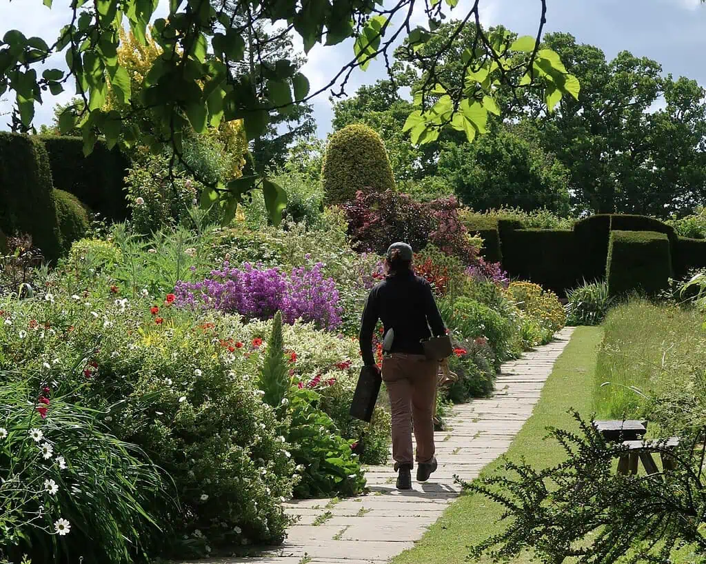 Een persoon met een opgerolde mat en tas loopt over een stenen pad door tuinen vol kleurrijke bloemen en gesnoeide heggen, genietend van de weelderige schoonheid van deze tuinen op een zonnige dag.
