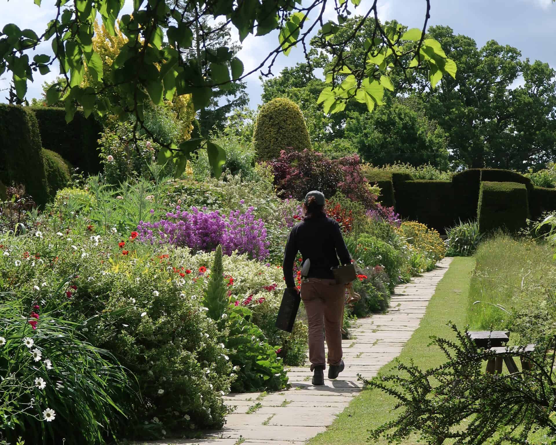 Een persoon met een opgerolde mat en tas loopt over een stenen pad door tuinen vol kleurrijke bloemen en gesnoeide heggen, genietend van de weelderige schoonheid van deze tuinen op een zonnige dag.