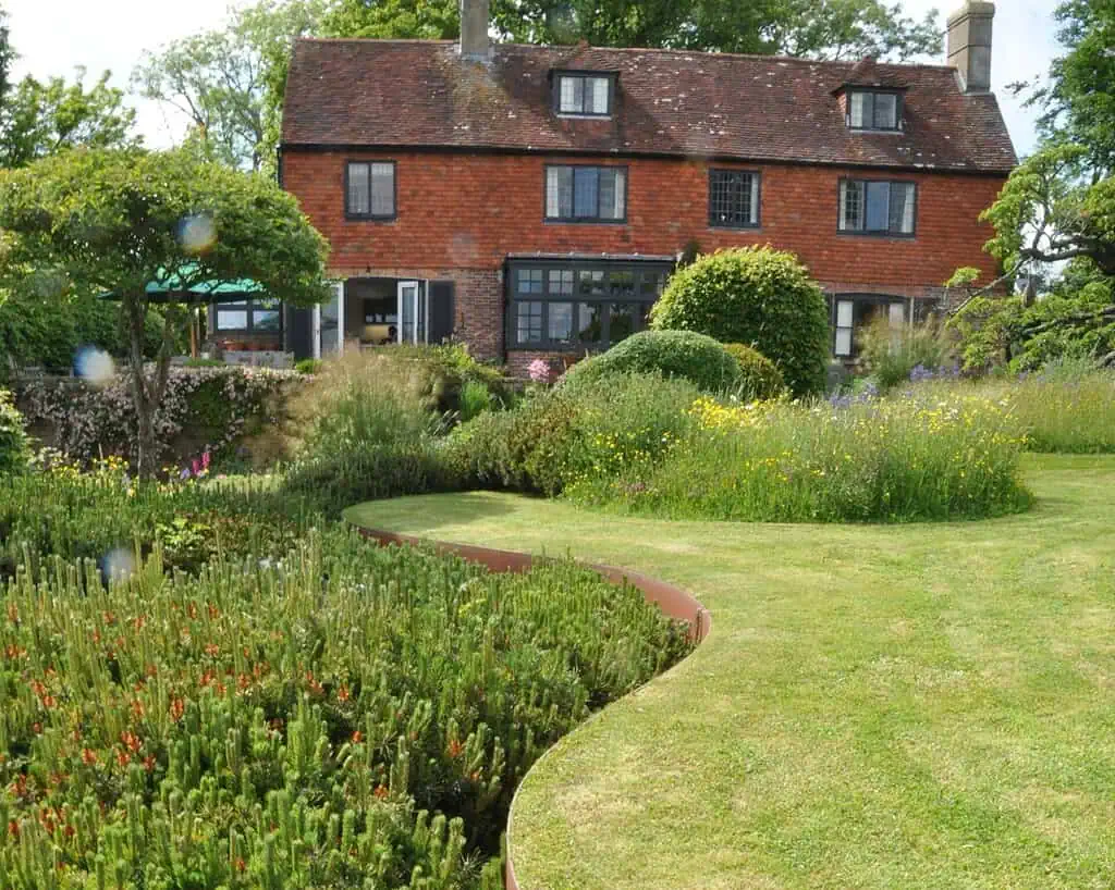 Een bakstenen huis met een rood pannendak staat achter een weelderige, goed onderhouden tuin geïnspireerd op de tuinen van de toekomst, met gebogen gazons, dicht groen, wilde bloemen en verschillende struiken onder een gedeeltelijk bewolkte hemel.