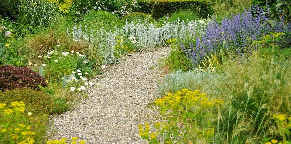 Een grindpad slingert door een weelderige tuin met Tuinenpracht met verschillende groene planten, gele bloemen, witte bloemen en paarse bloesems, die doen denken aan de betoverende tuinen in Duitsland.