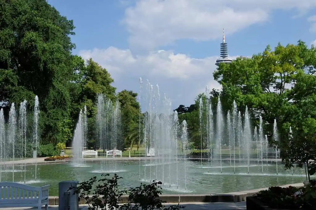 Een ronde waterfontein spuit meerdere stromen omhoog in een park omringd door weelderige groene bomen. Tuinenpracht in het hart van Duitsland, met witte bankjes en een hoge toren zichtbaar onder een gedeeltelijk bewolkte hemel.