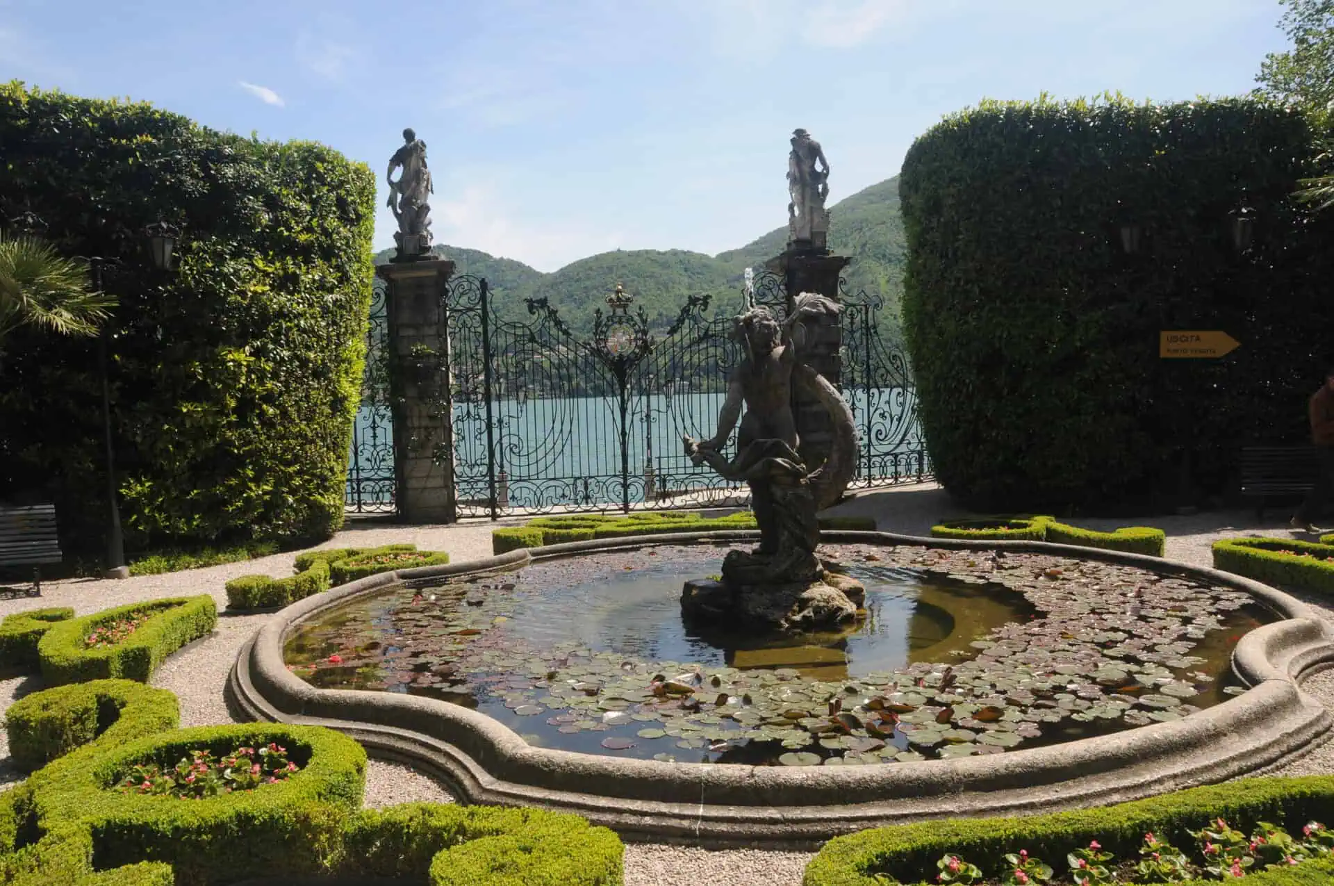 Een stenen fontein met een standbeeld omringd door waterlelies ligt in de goed onderhouden tuinen van Villa Carlotta, met gesnoeide heggen, grindpaden en een sierlijke ijzeren poort met uitzicht op een meer en groene heuvels op de achtergrond.
