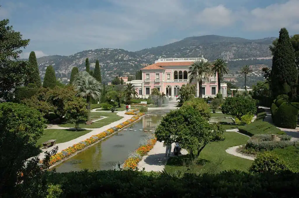 Een grote roze villa, geïnspireerd op Villa Ephrussi de Rothschild, staat tussen weelderige tuinen, palmbomen en gemanicuurde heggen. Een lang spiegelend zwembad met kleurrijke bloemen leidt naar het gebouw, met heuvels op de achtergrond.
