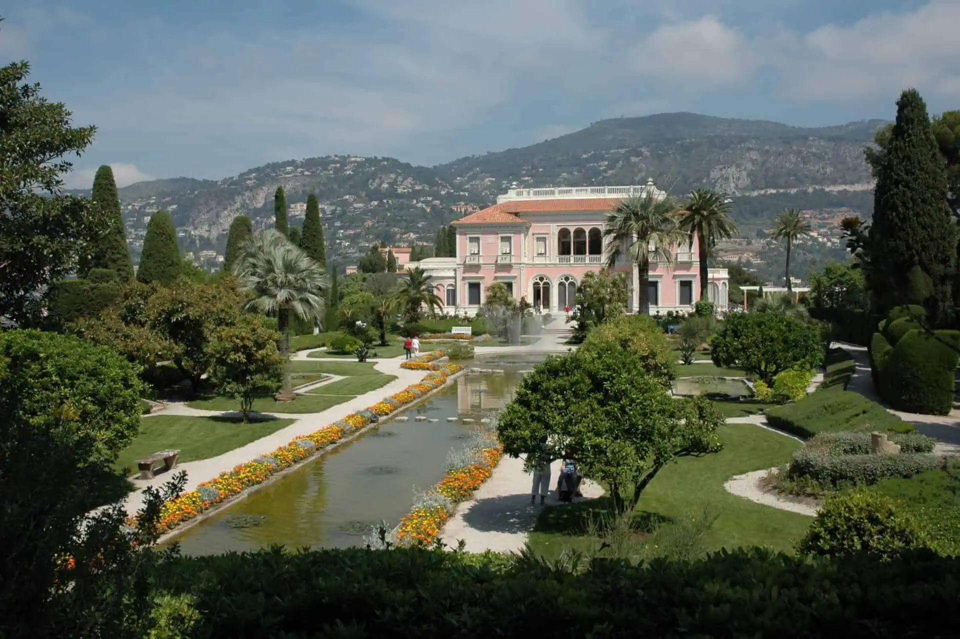 Een grote roze villa, geïnspireerd op Villa Ephrussi de Rothschild, staat tussen weelderige tuinen, palmbomen en gemanicuurde heggen. Een lang spiegelend zwembad met kleurrijke bloemen leidt naar het gebouw, met heuvels op de achtergrond.