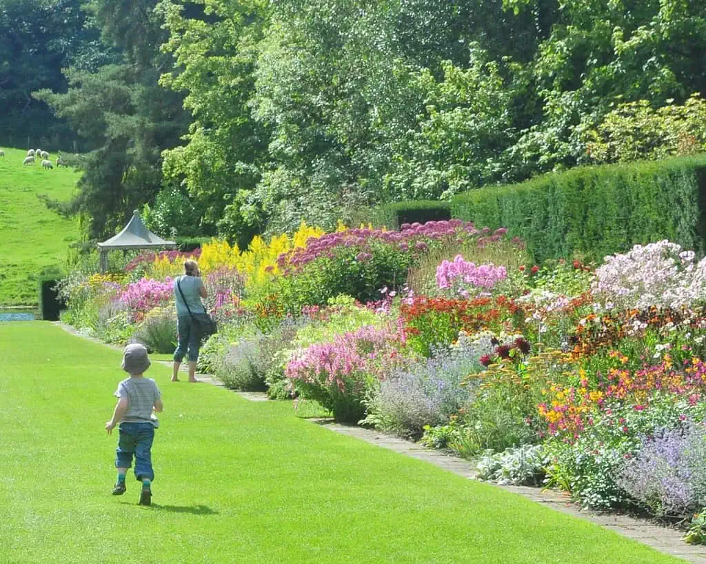 Een kind en een volwassene lopen over een pad langs een levendige Yorkshire tuin vol kleurrijke bloemen en groen op een zonnige dag. De scène roept de tijdloze landschappen op die de zussen Brontë koesterden.