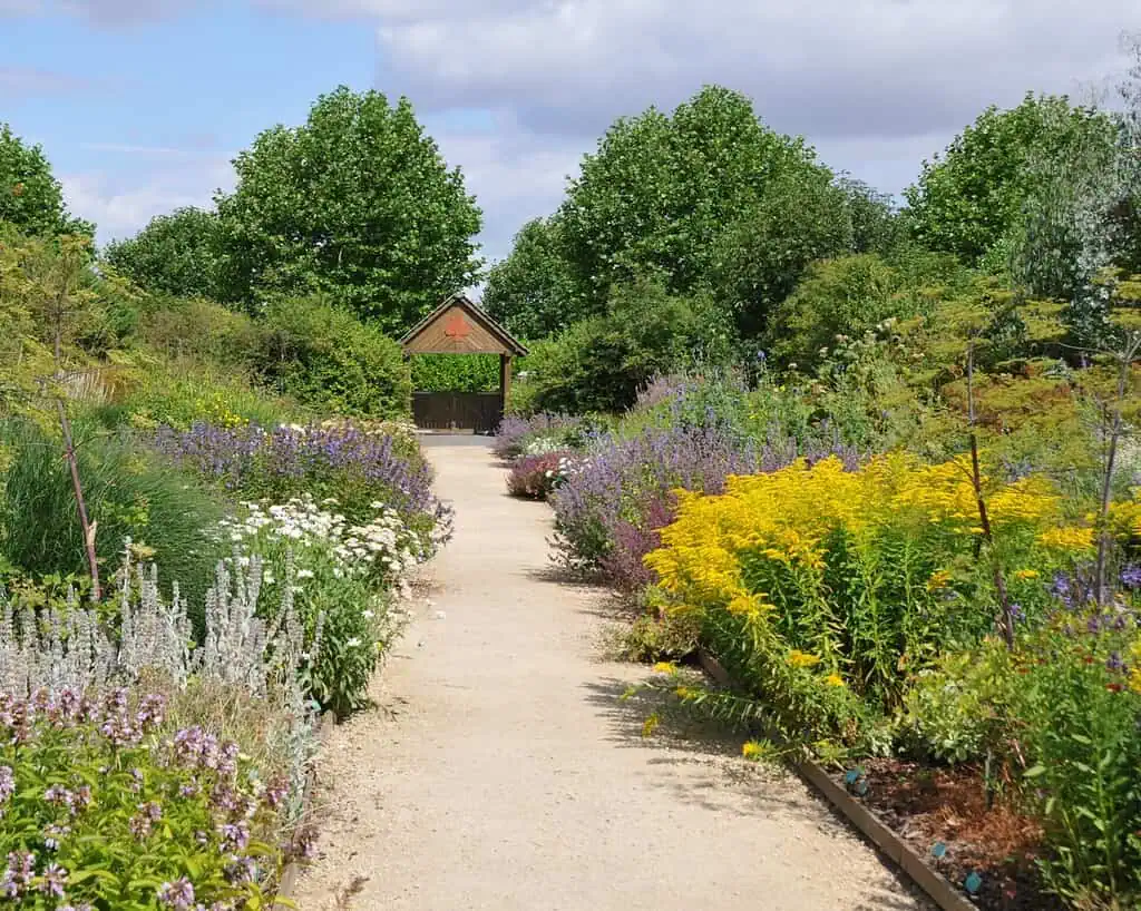 Een grindpad met kleurrijke bloemen en groene struiken leidt naar een kleine houten schuilplaats, die doet denken aan het platteland van Yorkshire waar de zussen Brontë zo van hielden, omringd door hoge bomen onder een halfbewolkte hemel.