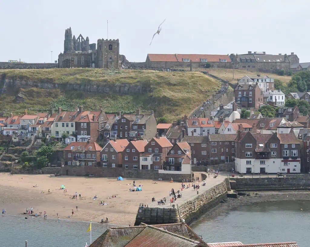 Een kustplaats in Yorkshire met mensen op een zandstrand, huizen met rode daken, een stenen pier en een grote ruïne van een abdij op een met gras begroeide heuvel op de achtergrond; een zeemeeuw vliegt over, wat doet denken aan scènes uit de romans van de zussen Brontë.