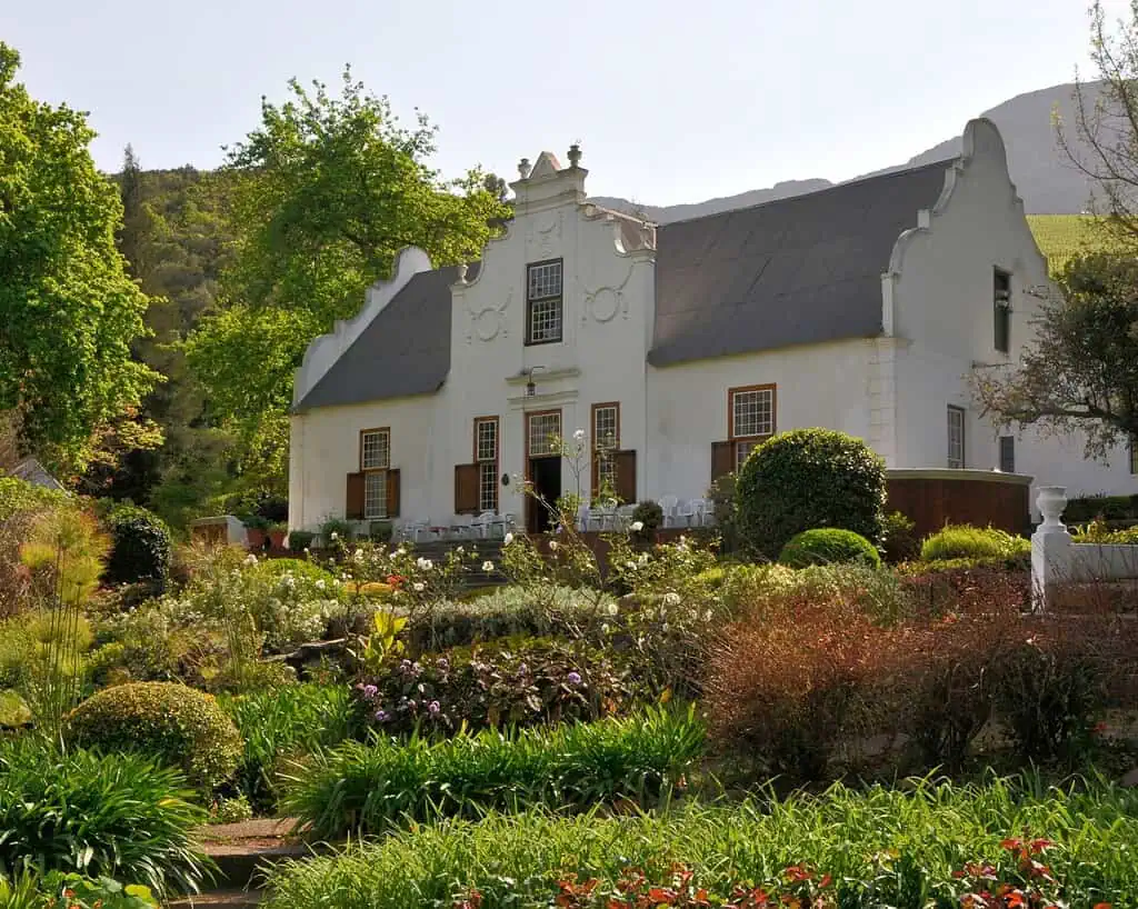 Een wit huis in Kaap-Hollandse stijl met sierlijke puntgevels staat te midden van weelderige, kleurrijke tuinen en groen, met heuvels en bomen op de achtergrond onder een heldere hemel.