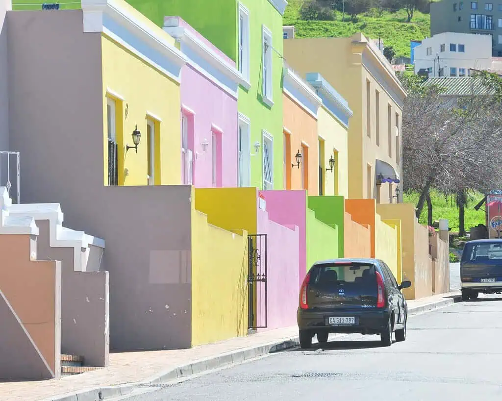 Een rij felgekleurde huizen omzoomt een zonnige straat, met een zwarte auto geparkeerd op de stoep. Elk huis is geschilderd in een andere levendige kleur, waaronder groen, geel, roze en oranje.