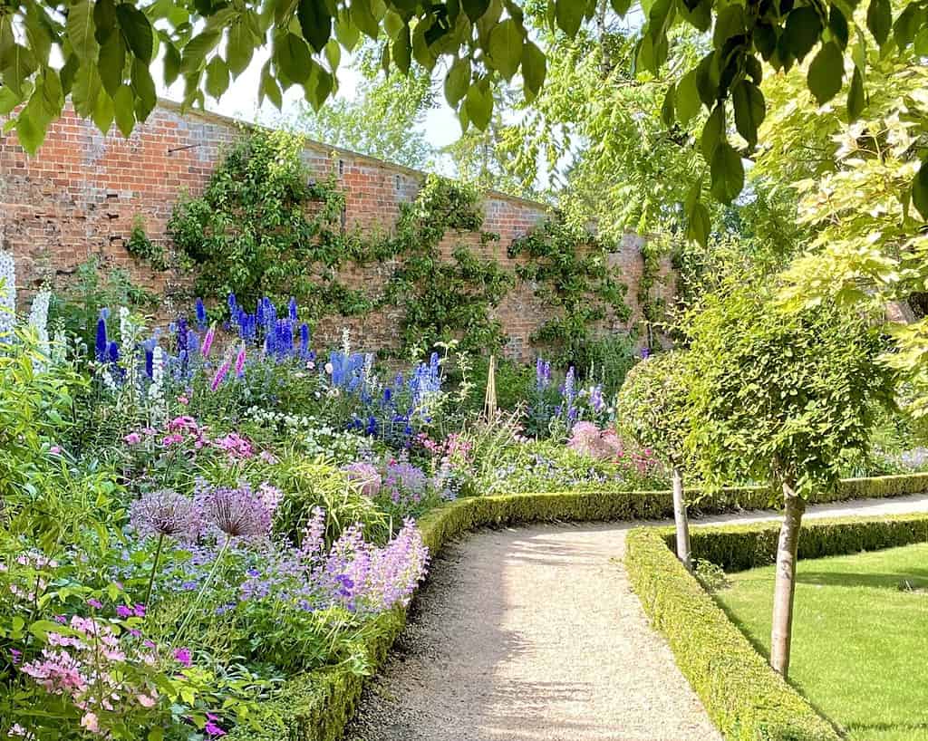 Een zonovergoten tuin in Zuid-Engeland heeft een grindpad dat kronkelt langs keurig gesnoeide heggen, kleurrijke bloeiende bloemen en hoge planten. Op de achtergrond bij Bath staat een bakstenen muur bedekt met groene wijnranken onder lommerrijke bomen.