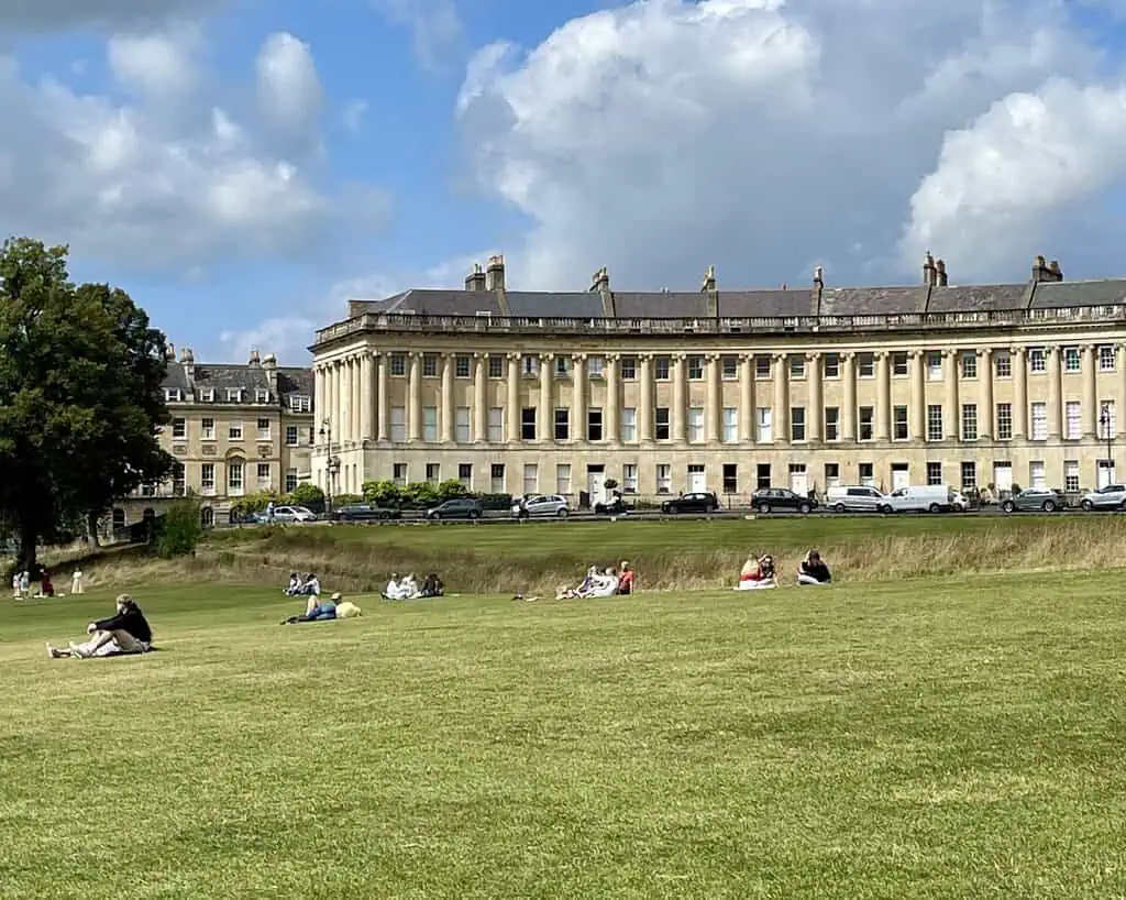 Mensen ontspannen op een groot grasveld voor de Royal Crescent, een lange gebogen rij historische Georgische gebouwen in Bath, Zuid-Engeland, onder een gedeeltelijk bewolkte hemel.