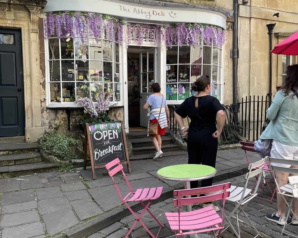 Een charmant café in Bath genaamd The Abbey Deli heeft roze stoelen, een groene tafel en buiten een bord met de tekst Open for Breakfast. Paarse bloemen hangen boven de deur en weerspiegelen de charme van Zuid-Engeland als mensen binnenkomen en buiten gaan staan.