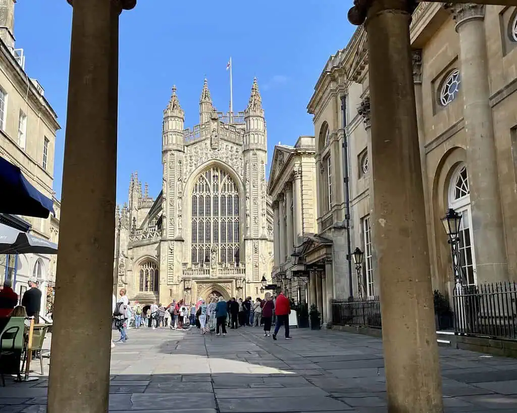 Mensen verzamelen zich buiten Bath Abbey op een zonnige dag in Zuid-Engeland, omlijst door stenen zuilen. De historische kathedraal heeft sierlijke gebrandschilderde ramen en hoge gotische torens tegen een strakblauwe lucht.