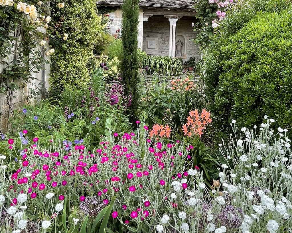 Een weelderige tuin in Zuid-Engeland vol kleurrijke bloemen en groen - felroze, oranje, paarse en witte bloemen - met een stenen gebouw en een gewelfde nis met een standbeeld op de achtergrond.