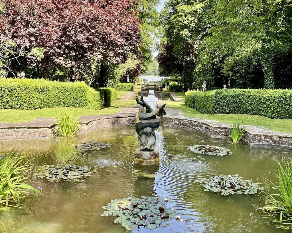 Een tuinvijver in Zuid-Engeland met waterlelies is voorzien van een gebeeldhouwde fontein van een jongen die een vis vasthoudt. De vijver wordt omzoomd door verzorgde hagen, levendige bomen en een stenen pad dat doet denken aan charmante Bathse tuinen.