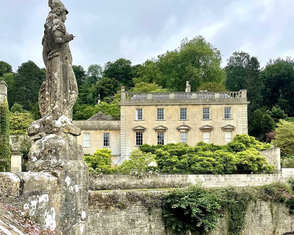Een groot stenen herenhuis met hoge ramen en weelderig groen aan de voorkant, gedeeltelijk verborgen door een oud stenen standbeeld en een met mos bedekte muur op de voorgrond; dichte bomen vullen de achtergrond onder een bewolkte Zuid-Engeland lucht.