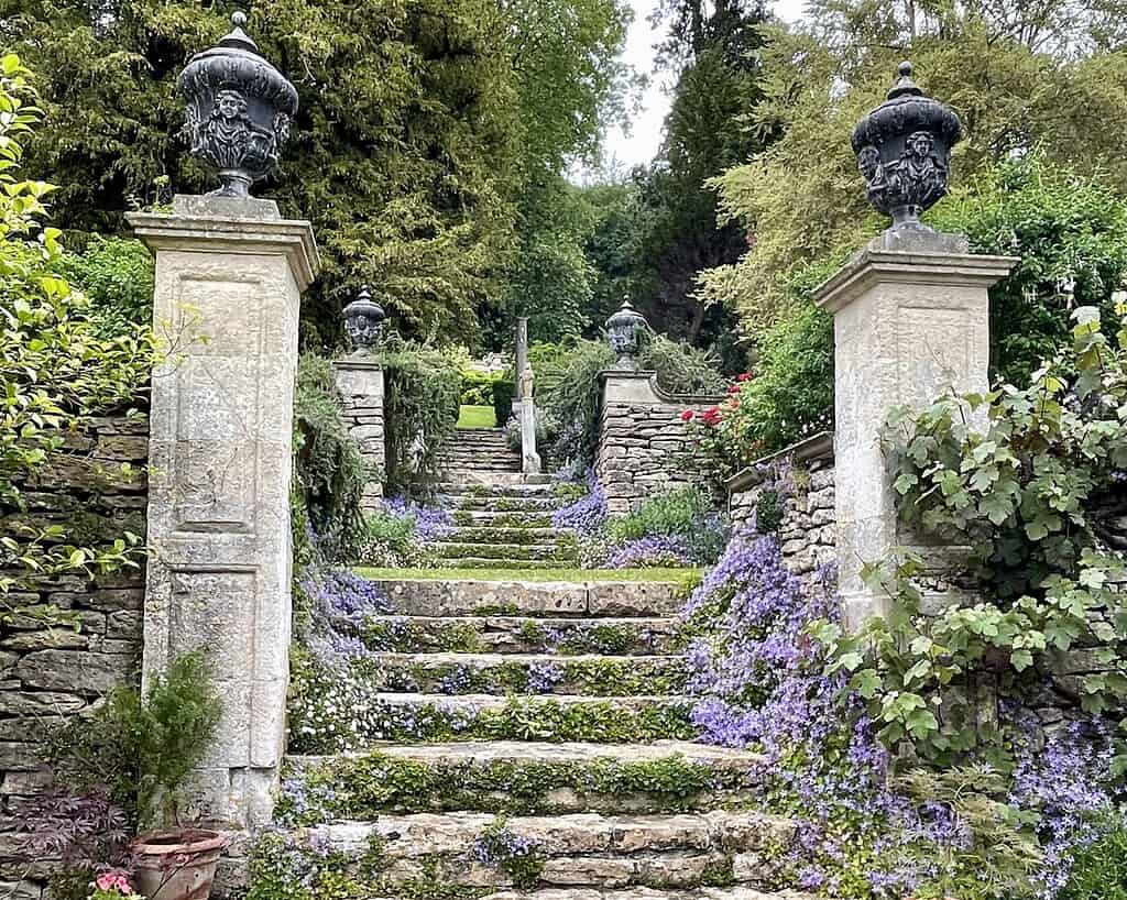Stenen trappen omzoomd door stenen muren en pilaren met sierlijke urnen, bedekt met kruipende paarse bloemen en groen, leiden omhoog door een weelderige, aangelegde tuin die doet denken aan Bath in Zuid-Engeland, omringd door hoge bomen.