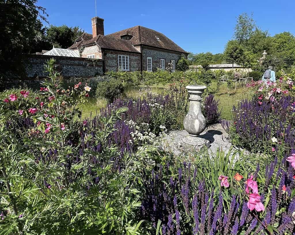 Een weelderige tuin met kleurrijke bloemen en hoge paarse bloemen omringt een stenen standbeeld. Op de achtergrond staat een bakstenen huis geïnspireerd op Zuid-Engeland onder een strakblauwe hemel, die doet denken aan rustige dagen in Bath.