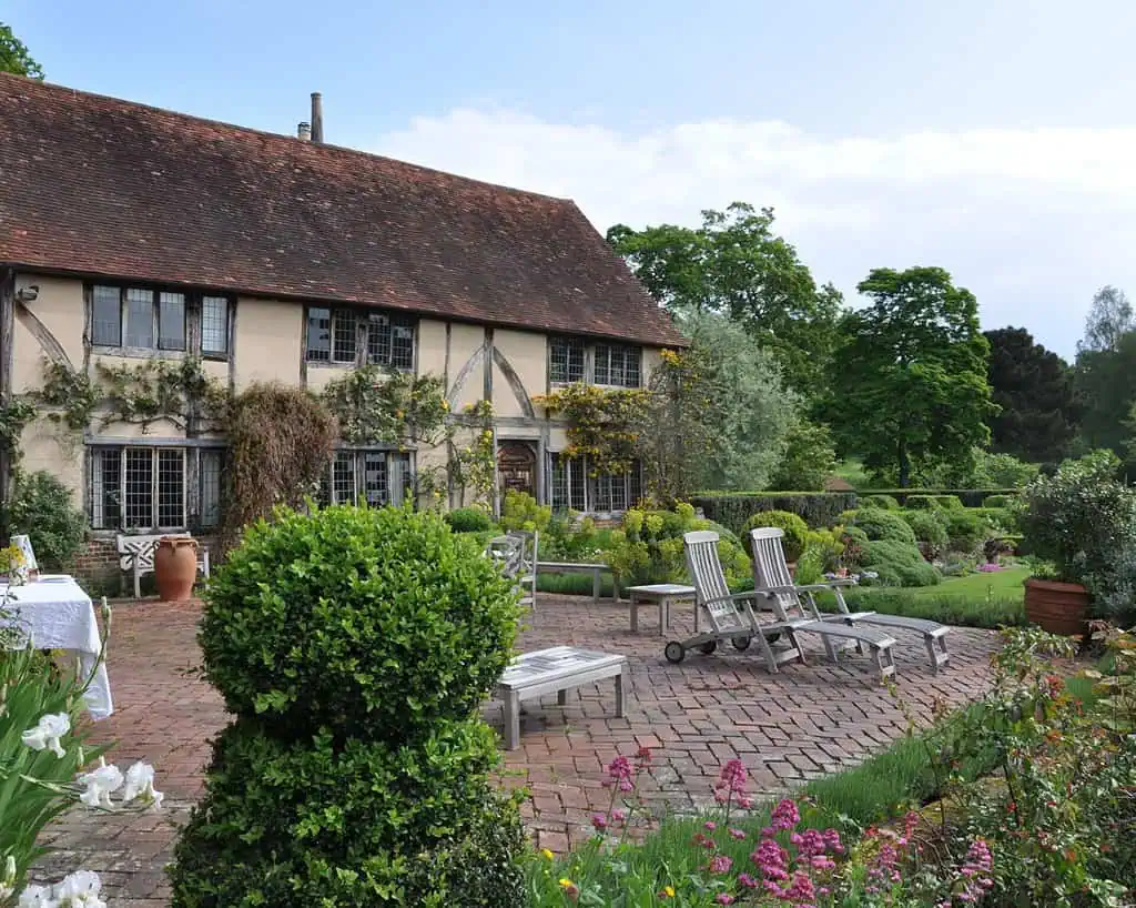 Een witte metalen kan met een afbeelding van een haan houdt groen en geel gebladerte vast op een houten dienblad. Daarachter wijst een bord met de tekst "WEST COURT LODGE" op de charmante vondsten van Antiekreis Ardingly.