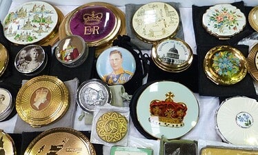 Een charmante verzameling antieke decoratieve poederdozen en compacts uit Antiekreis Ardingly, met koninklijke portretten, kronen, bloemmotieven en kaarten, allemaal netjes gerangschikt op een display.