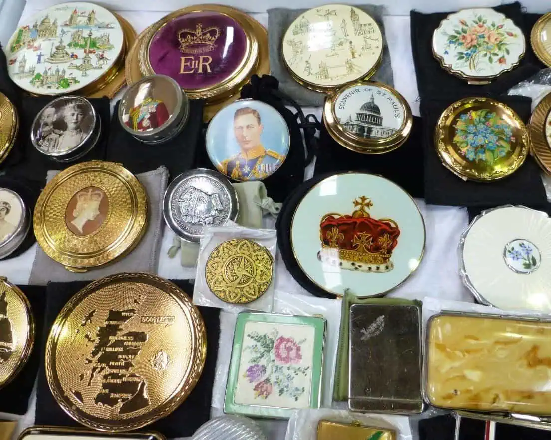 Een charmante verzameling antieke decoratieve poederdozen en compacts uit Antiekreis Ardingly, met koninklijke portretten, kronen, bloemmotieven en kaarten, allemaal netjes gerangschikt op een display.